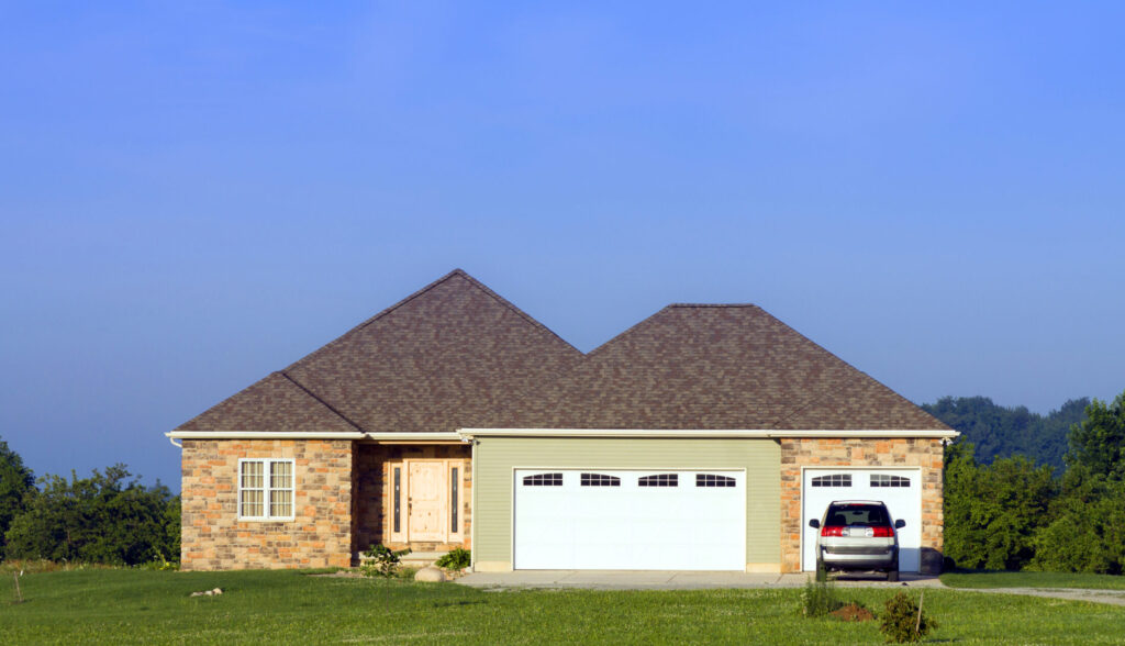 Single-story stone and siding ranch-style house with an attached three-car garage and a car parked in the driveway against an expansive green lawn and blue sky